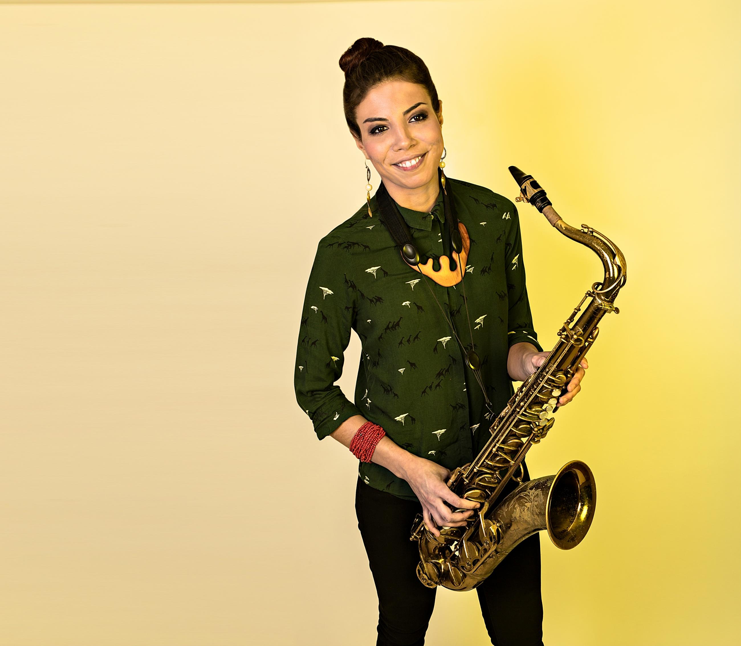 Melissa Aldana standing in front of yellow wall holding tenor saxophone
