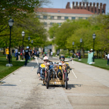 Two students pedal a go-kart made of two bicycles on a sidewalk on a sunny day.
