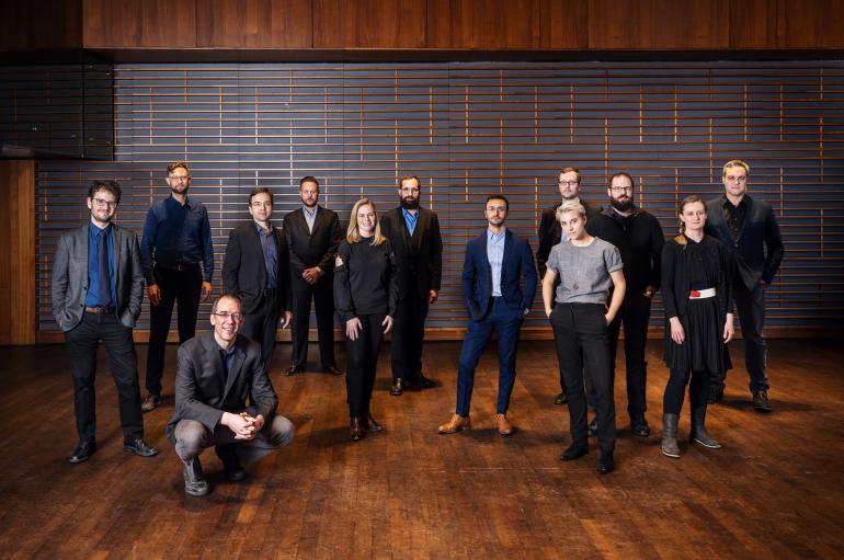 The Grossman Ensemble members standing at various distances in a large room with dark brown walls and wood floors