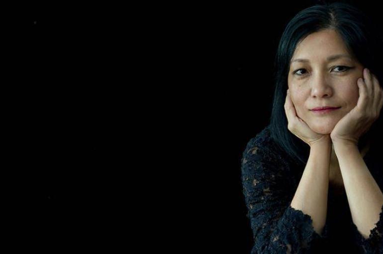 Sylvia Wang wearing black in front of a black background, cupping her chin in her hands