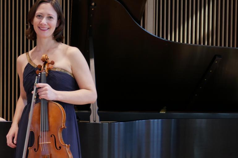 Rose Wollman wearing a blue dress and holding a viola in front of a piano