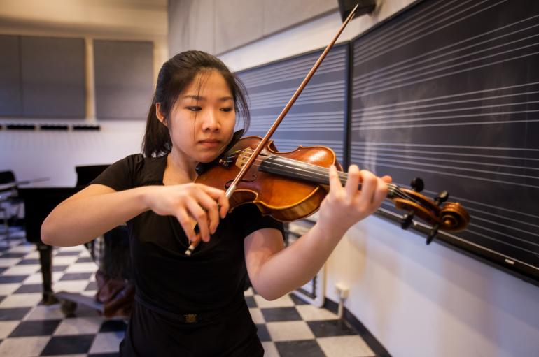 A student practices violin in front of a black board with white staff lines and a grand piano