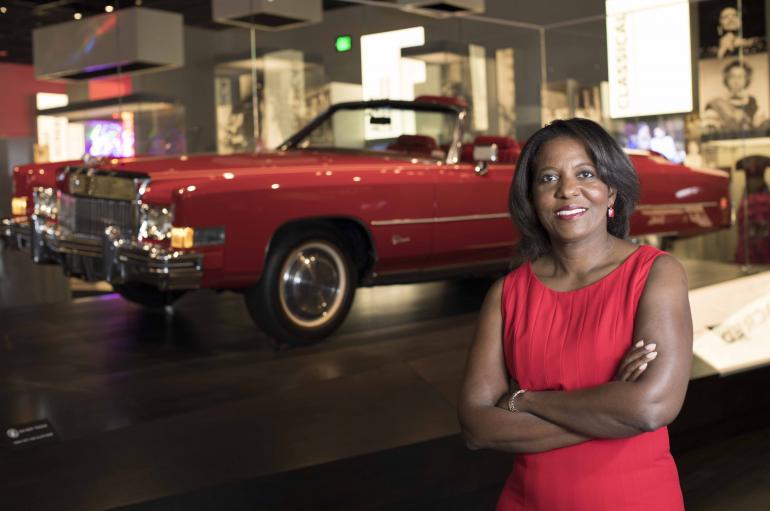 Dwandalyn R. Reece, a black woman wearing a read dress, stands in front of a red vintage hot rod