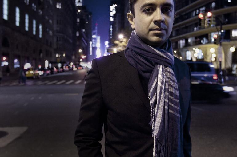 Vijay Iyer standards wearing a dark blazer and striped scarf in a dark street in New York