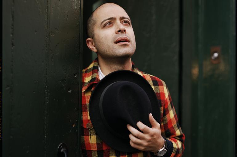 Mahan Esfahani shown wearing a red, brown, and yellow plaid jacket and holding brown hat to his chest