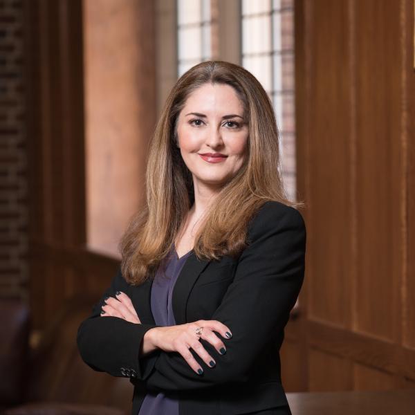 Photo of a woman in a black blazer and grey blouse with long brownish hair.